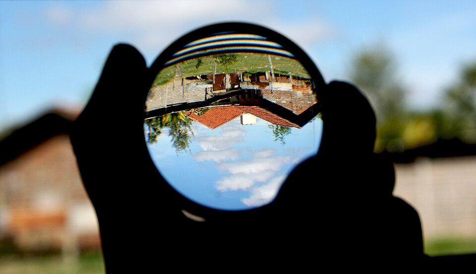 A convex glass lens held up against a rural scene, the converged light forming a sharp upside-down image of the houses and sky inside the lens.