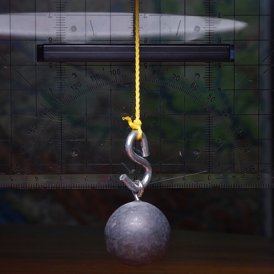 A heavy metal pendulum bob hanging motionless on a yellow cord in front of a laboratory protractor scale, forming a simple pendulum.