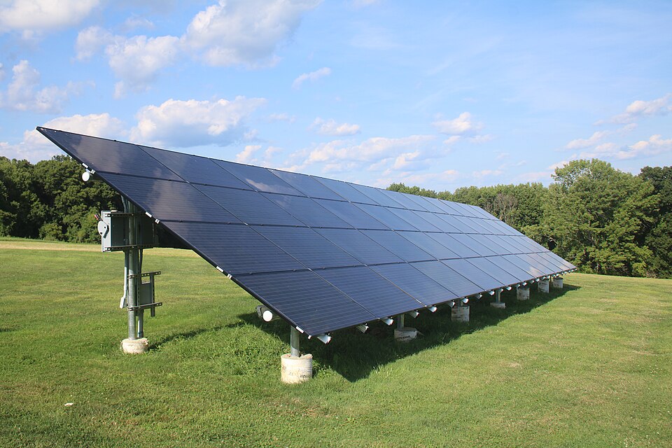 A ground-mounted solar photovoltaic array tilted towards the sky in a grass field, its dark modules made up of a visible grid of individual solar cells.