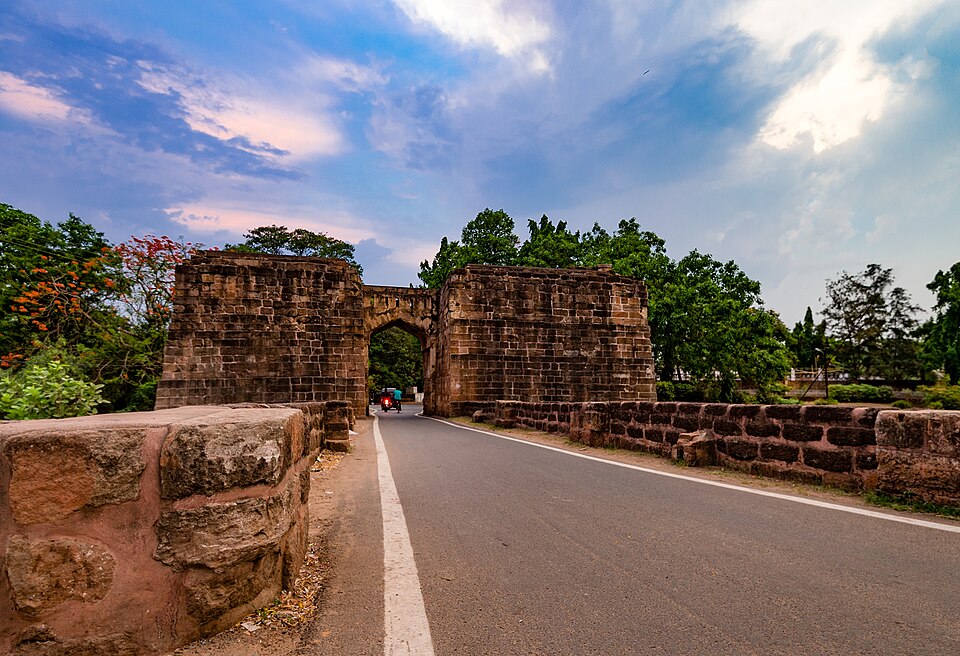 The ruined stone gateway of Barabati Fort at Cuttack, Odisha, its heavy laterite blockwork flanking a road that runs through the arch across the old moat causeway.
