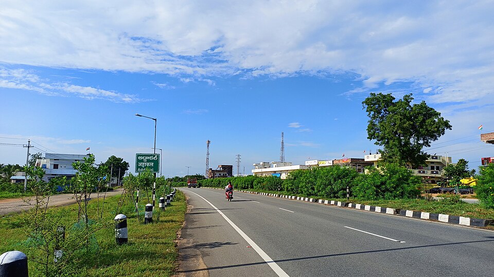 A divided stretch of National Highway 44 running past Addakal in Telangana, with a green highway signboard and painted kerb markers along the carriageway.