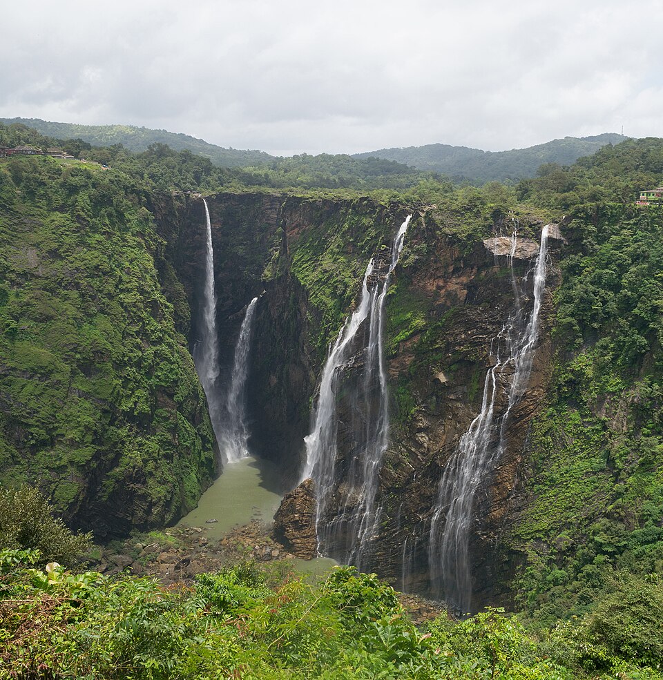 Four separate white cascades of water plunge side by side down a steep forested gorge into a green pool far below, with the wooded hills of the Western Ghats behind.