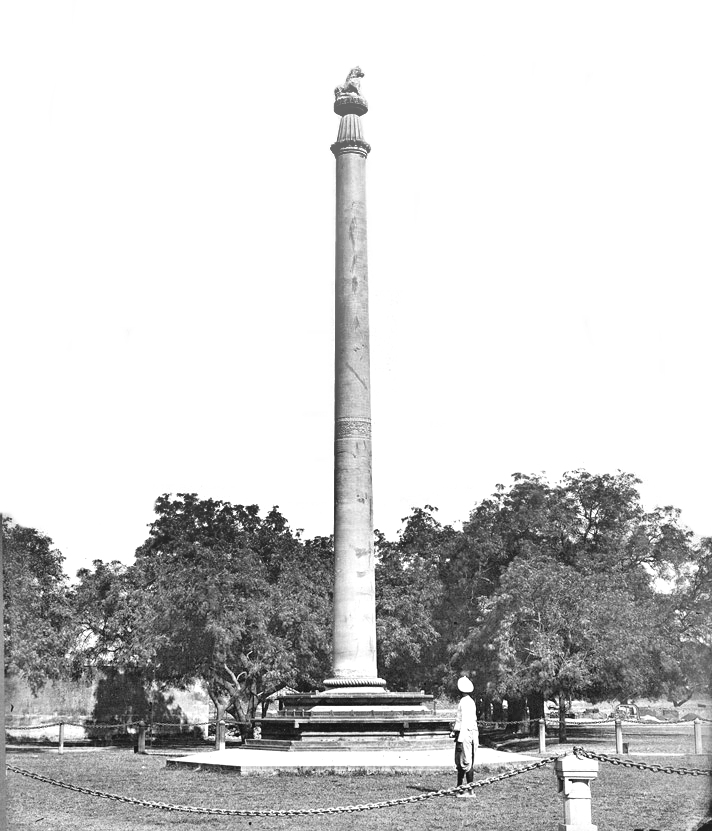 The tall polished monolithic Ashokan pillar standing on a plinth inside the grounds of Allahabad Fort, topped by a lion capital, photographed in the 1870s with a man standing beside it for scale.