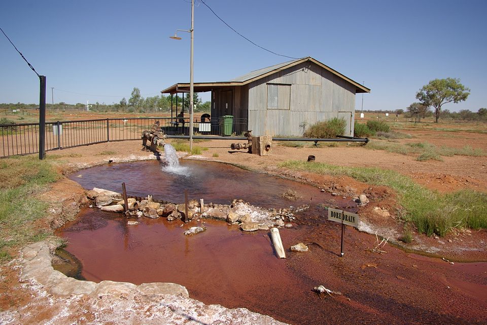 A free-flowing artesian bore at Thargomindah in outback Queensland, jetting water from a pipe into a rust-red bore drain beside a corrugated-iron shed under a clear sky.