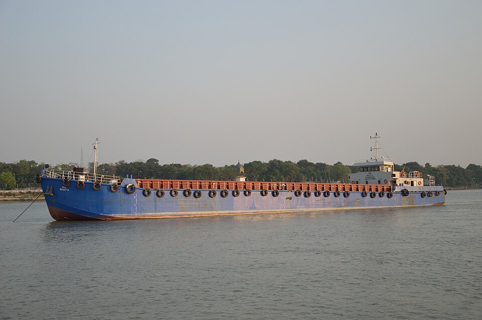 A long blue inland general-cargo vessel under way on the river Hooghly, the Kolkata reach of National Waterway 1, with the wooded riverbank behind it.