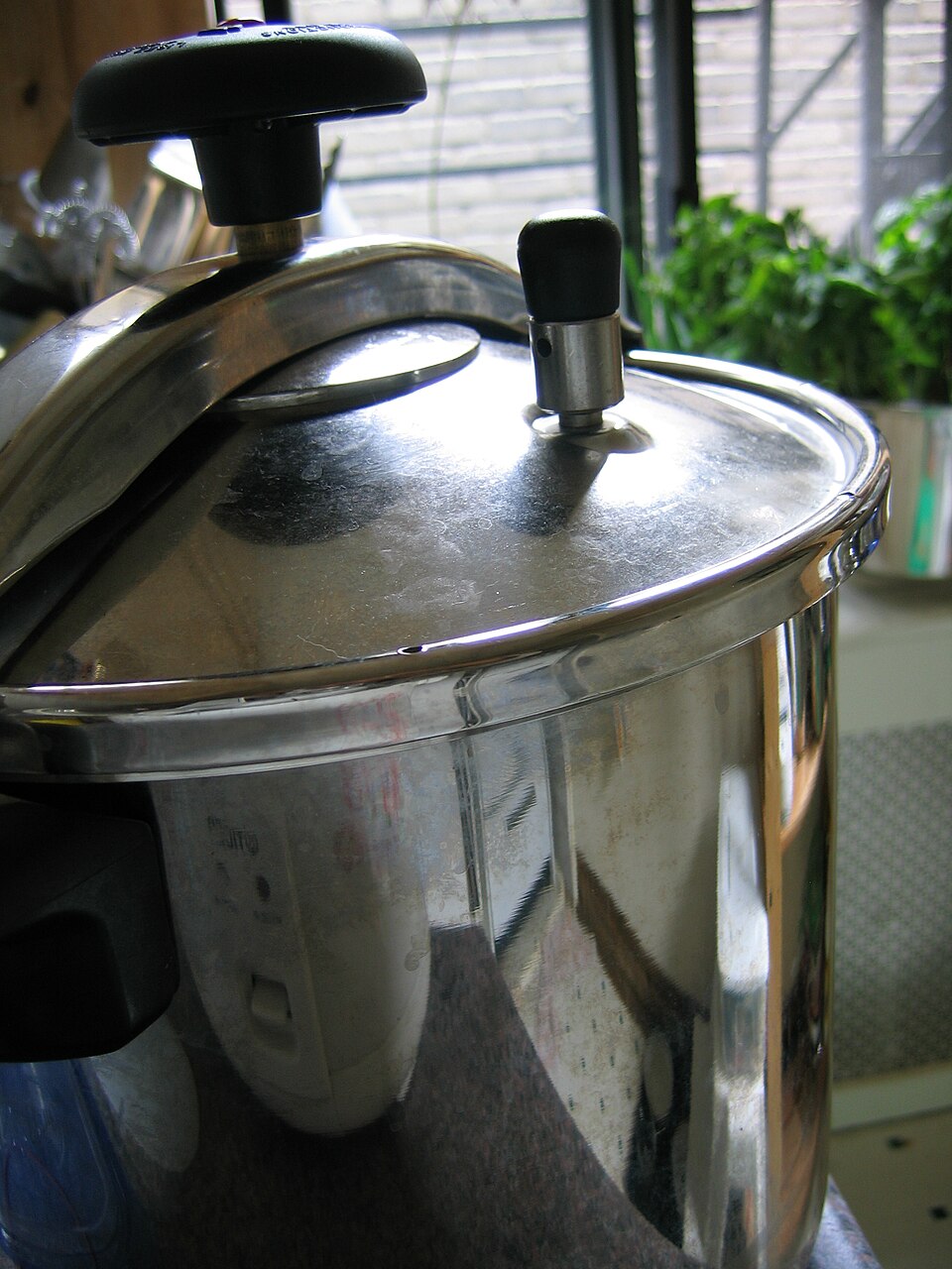 A stainless-steel domestic pressure cooker seen close up, with its locking lid clamped down and the pressure-regulating valve standing up on top of the lid.