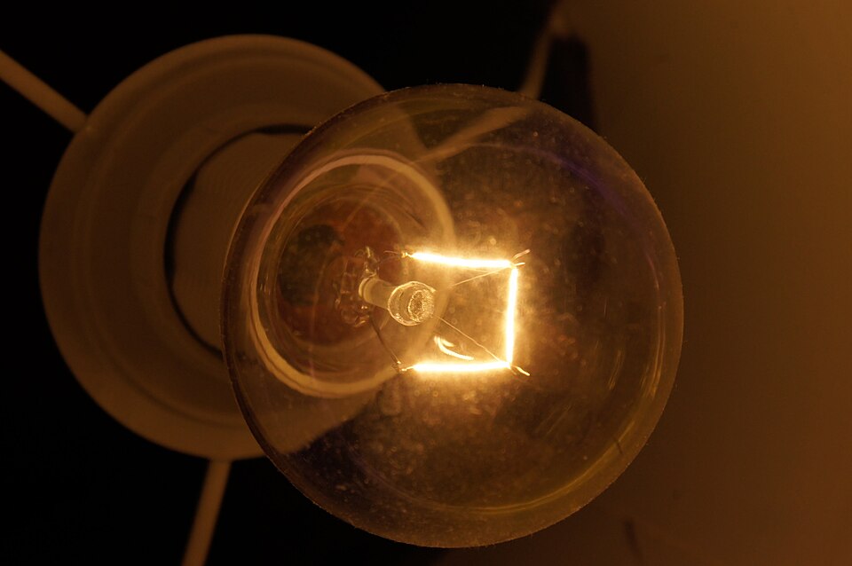 A lit clear incandescent light bulb in a ceiling holder, photographed close up so that the bright glowing tungsten filament inside the glass bulb is clearly visible.