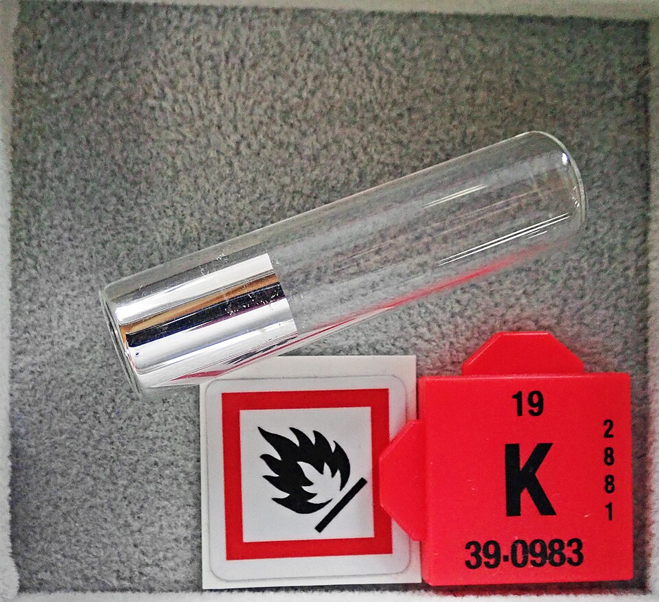 A bright silvery piece of potassium metal sealed inside a glass vial, lying beside a flammable-hazard label and a red periodic-table tile marked 19 K 39.0983.