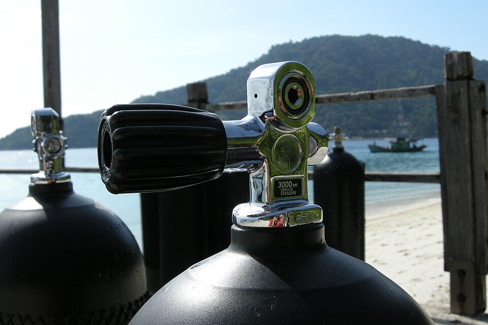 Black compressed-gas diving cylinders standing on a jetty by the sea, the nearest one showing its chrome pillar valve and handwheel stamped with a 3000 psi service pressure.