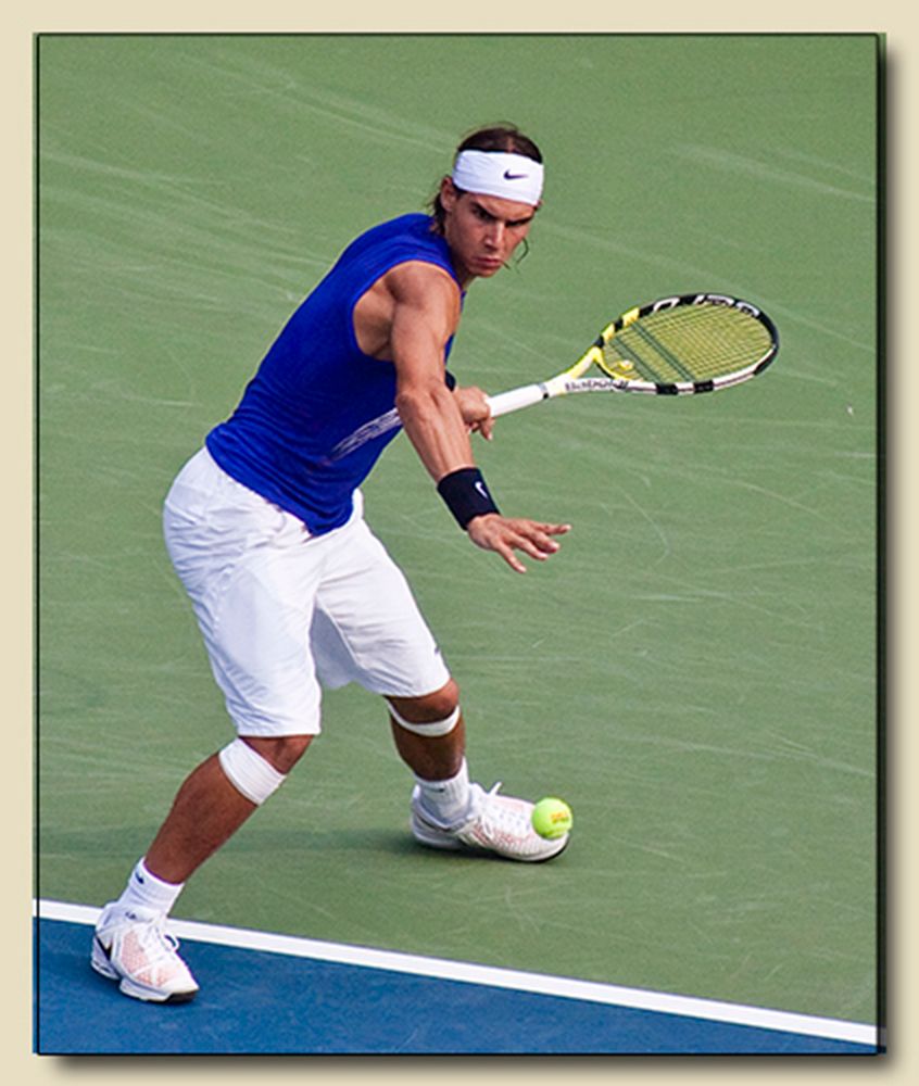 Rafael Nadal in a blue sleeveless shirt and white shorts, racquet extended, striking a low ball on a tennis court.