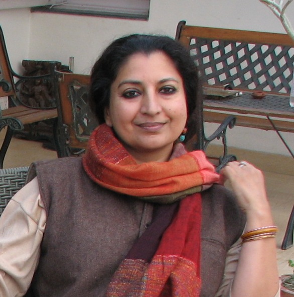 Portrait of the Hindi novelist Geetanjali Shree, seated outdoors in a brown jacket with a red and orange striped scarf.
