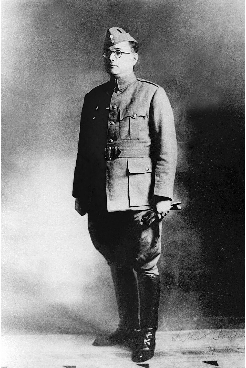 Full-length black-and-white photograph of Subhas Chandra Bose standing in Indian National Army uniform — side cap, round spectacles, belted tunic and riding boots — with his signature written across the lower right of the print.