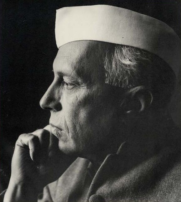 Black-and-white profile portrait of Jawaharlal Nehru wearing a white Gandhi cap, resting his chin on his hand.