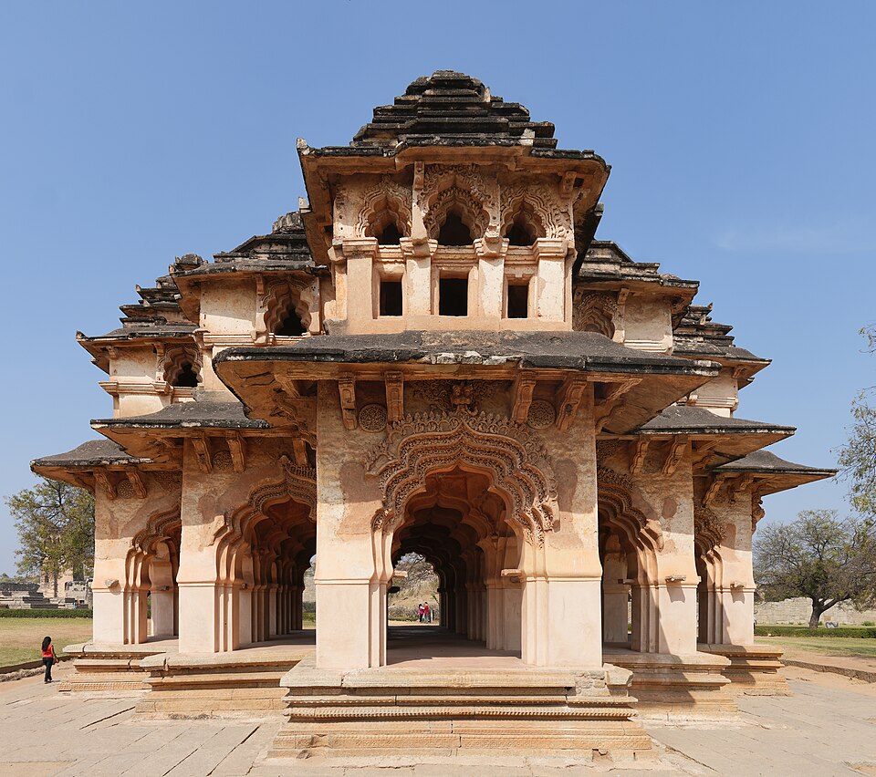 A frontal view of the two-storeyed Lotus Mahal at Hampi, with its row of lotus-bud arches, carved stucco decoration and tiered pyramidal roof towers rising against a clear sky.