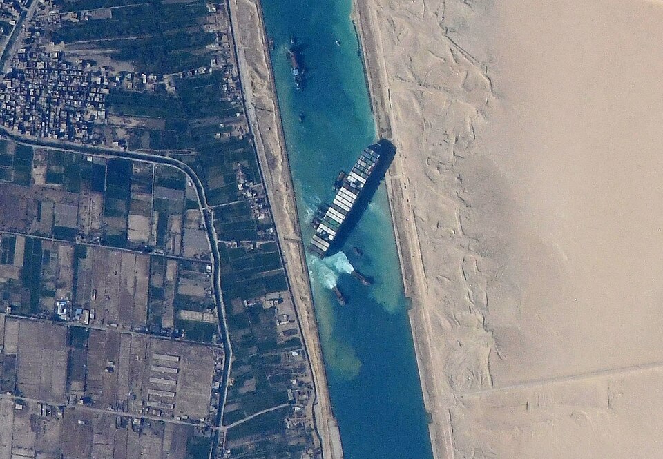 Satellite view of the Suez Canal, a dead-straight man-made channel cut between desert on one side and irrigated farmland on the other, with a large container ship lodged across it.
