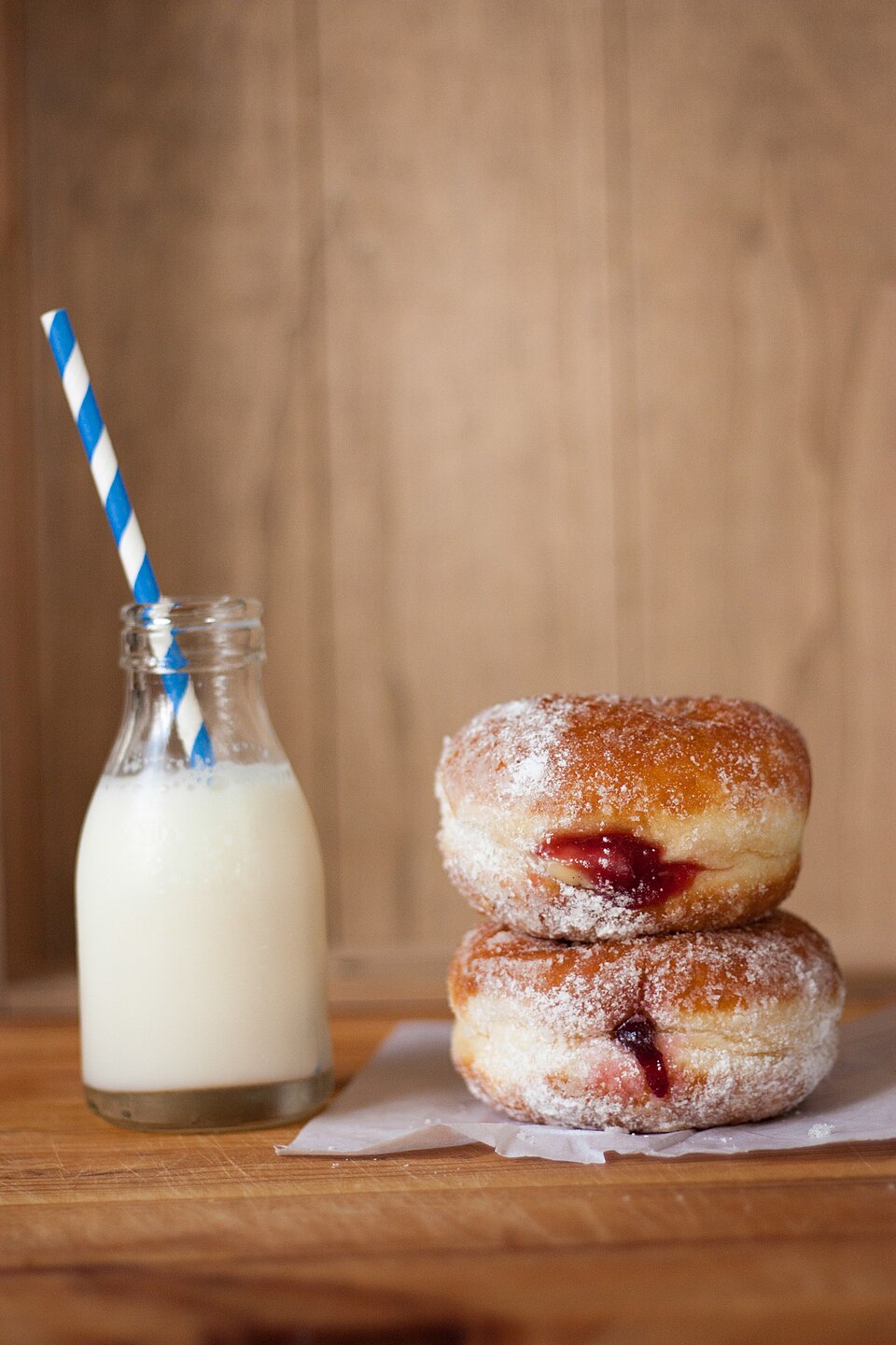 A small glass bottle of opaque white milk with a striped paper straw, standing on a wooden board beside two sugar-dusted jam doughnuts.
