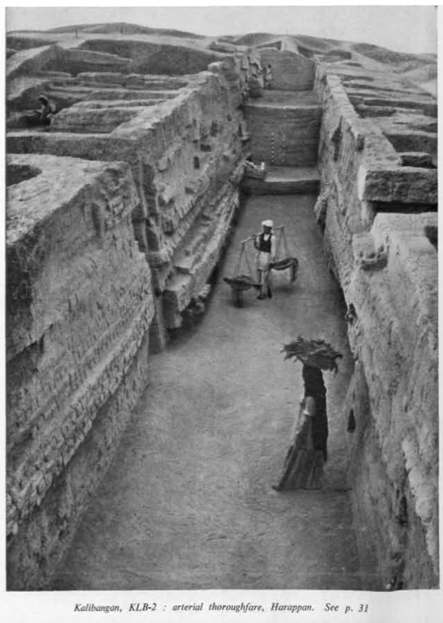 Archaeological Survey of India excavation photograph of Kalibangan, showing a deep excavated Harappan street running between mud-brick walls with two workers standing in it.