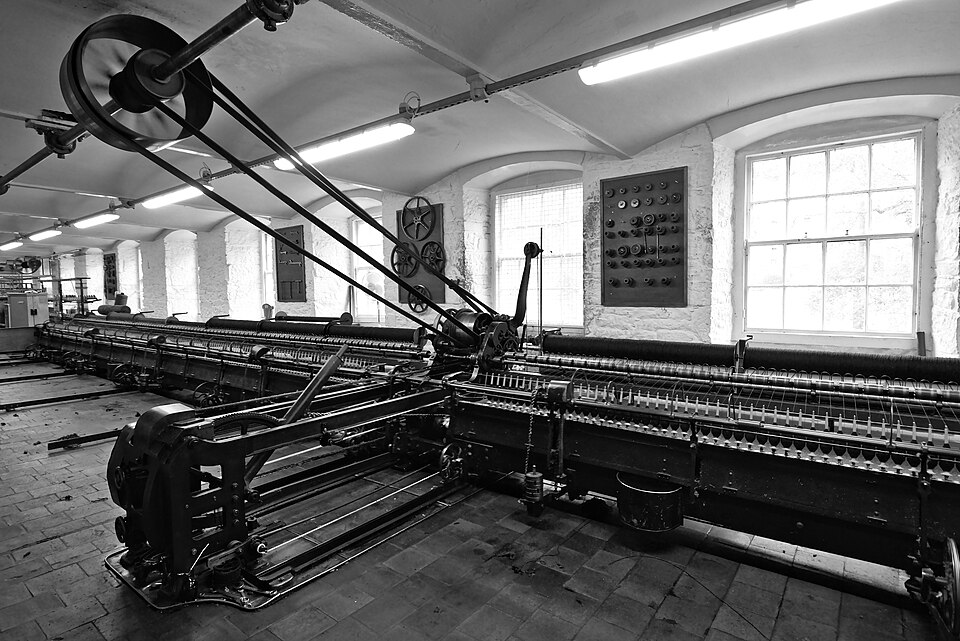 A preserved spinning mule in a mill room, its long carriage of spindles and rollers driven by an overhead belt and pulley.