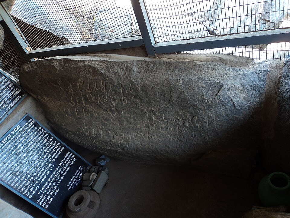 The Ashokan minor rock edict at Maski, a low granite boulder carved with lines of Brahmi script, protected behind a metal grille beside an Archaeological Survey information board.