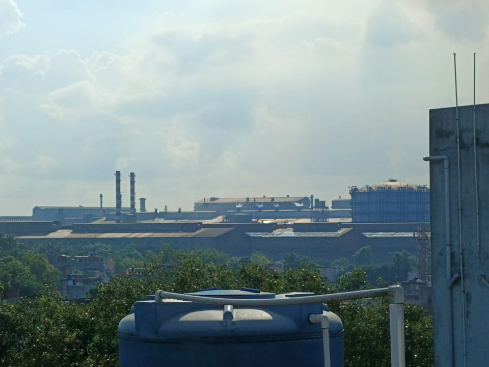 Rooftop view across Jamshedpur towards the Tata Steel works, showing long mill sheds, tall chimneys and a large gas holder above the tree line.