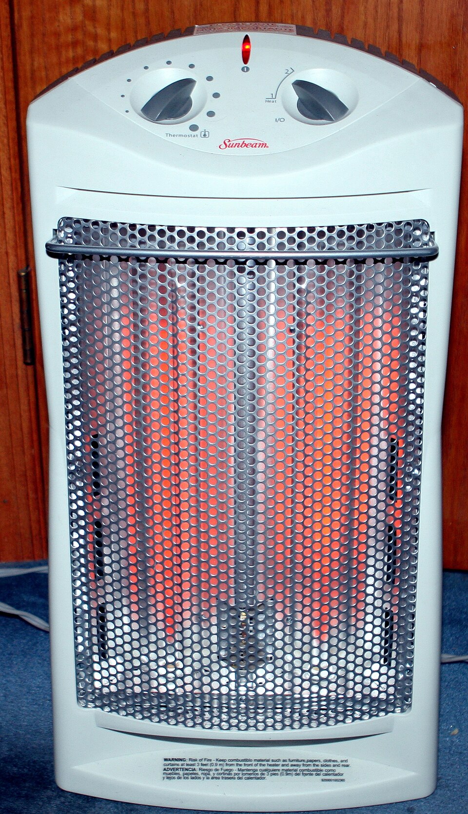 An upright domestic electric room heater switched on, its resistance heating elements glowing orange-red behind a perforated metal guard, with thermostat and heat-setting knobs on top.