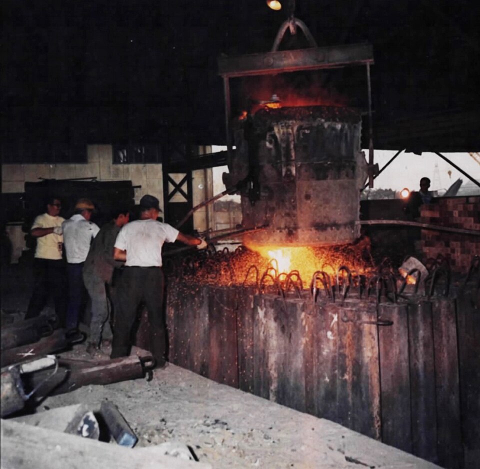 Steelworkers at a steel mill guide a glowing stream of molten steel from an overhead ladle into a row of casting moulds, showering sparks.