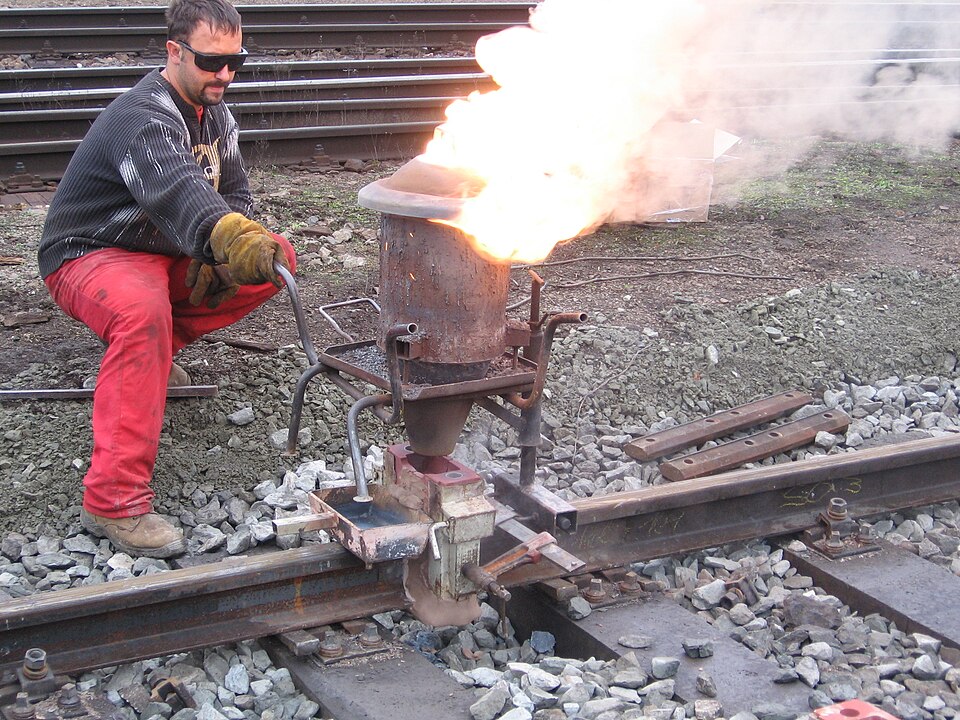 A track worker kneels beside a railway rail as a thermite crucible clamped over the rail joint flares with a tall white flame, welding the two rail ends together.