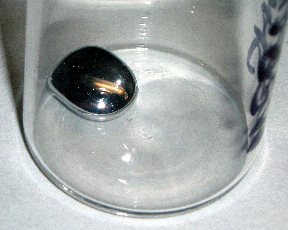 A single bead of liquid mercury, silvery and mirror-bright, resting in the bottom of a clear glass vessel at room temperature.