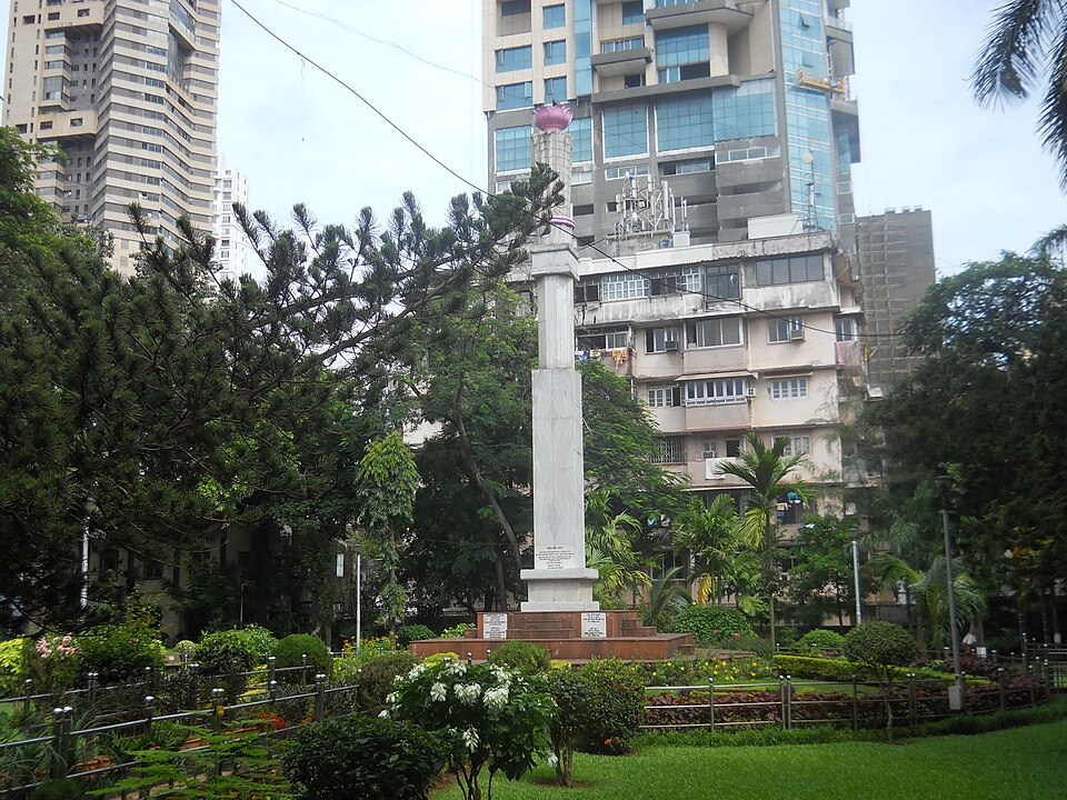 The tall white memorial pillar with inscribed plaques at its base standing in the landscaped grounds of August Kranti Maidan, Mumbai, with city high-rise buildings behind it.