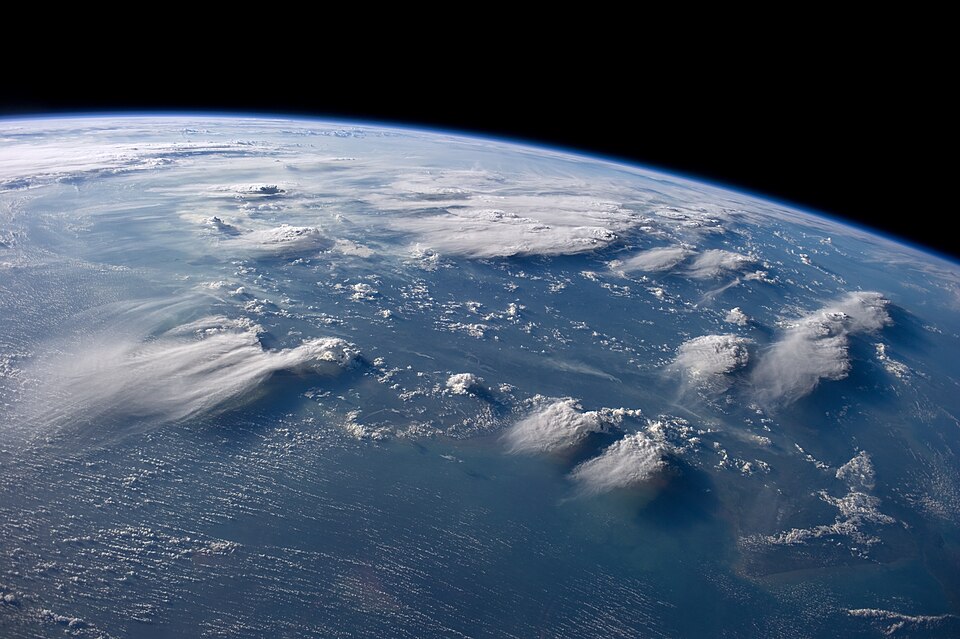 A view from the International Space Station of towering cumulonimbus thunderheads with spreading anvil tops rising over the sea near Borneo, confined within the thin blue band of atmosphere visible at the Earth's edge.
