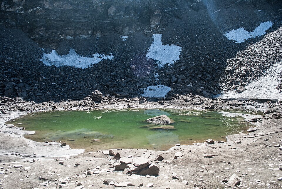 The small shallow green-water tarn of Roopkund lying in a bowl of bare rock and scree, with patches of snow on the slopes above it.