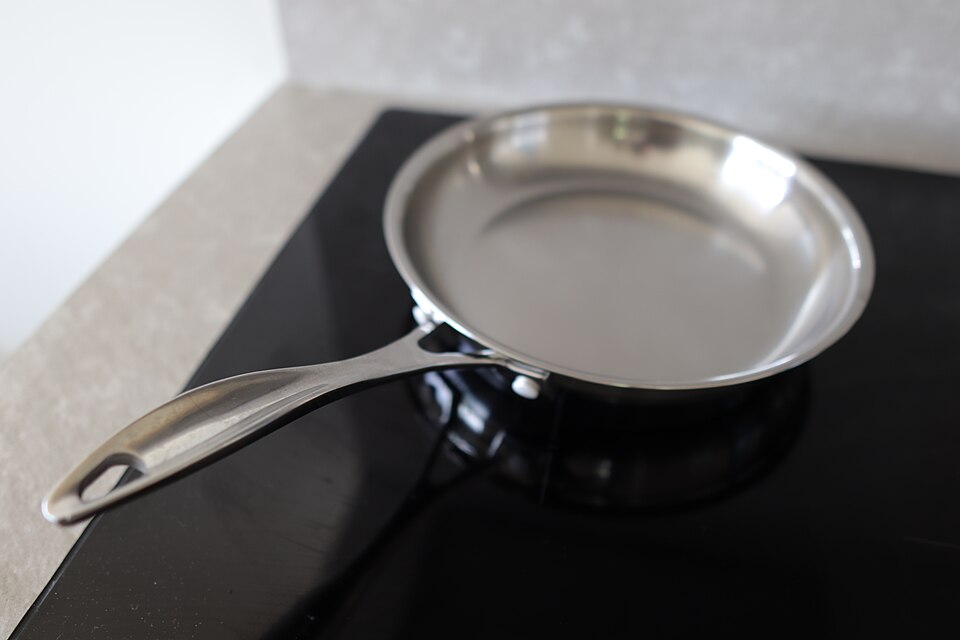 A clean, unmarked stainless steel frying pan with a bright mirror-like interior and a steel handle, resting on a black hob.