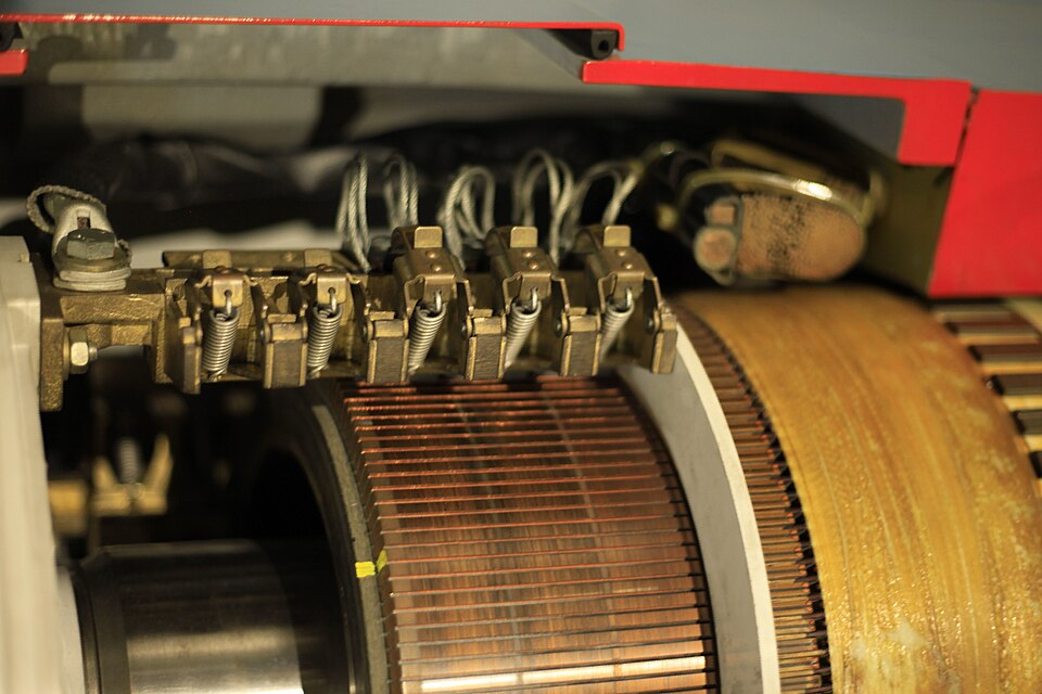 Close-up of the commutator and spring-loaded carbon brushes of a large DC electric motor, with copper armature windings visible