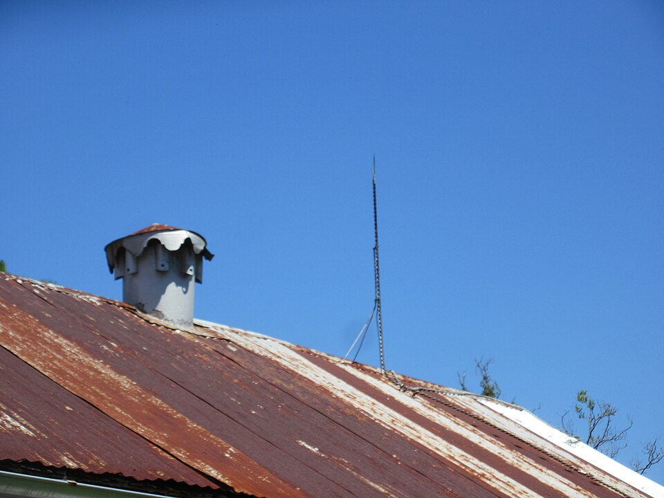 A tall pointed metal lightning conductor rod mounted on a building's rooftop against a clear blue sky, beside a chimney