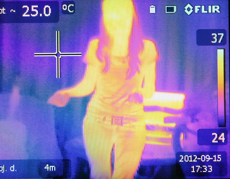 An infrared thermographic image of a standing person in false colour, the warm body glowing yellow-orange against a cooler blue-purple background, with a temperature scale
