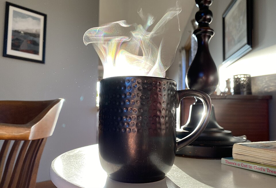 A black mug of hot coffee with steam rising visibly in the light as it cools on a table