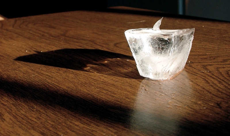 A melting ice cube on a wooden table with a pool of water beneath it