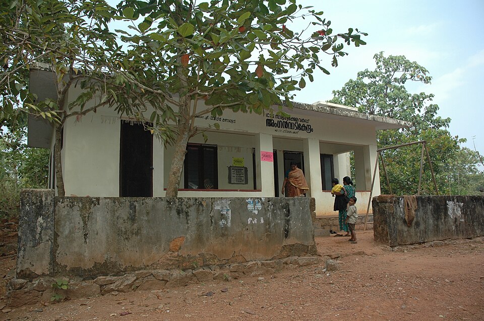 A small whitewashed village anganwadi building with an open verandah and Malayalam signage above the entrance, a tree in front and a few women and children standing near the doorway.