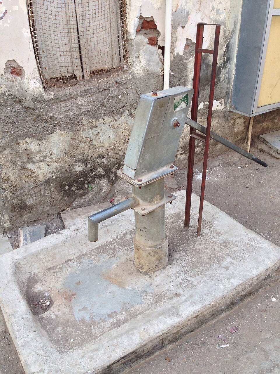 A metal India Mark II hand pump with a long lever handle mounted on a raised concrete platform against a weathered wall, with a spout over a shallow drainage apron.