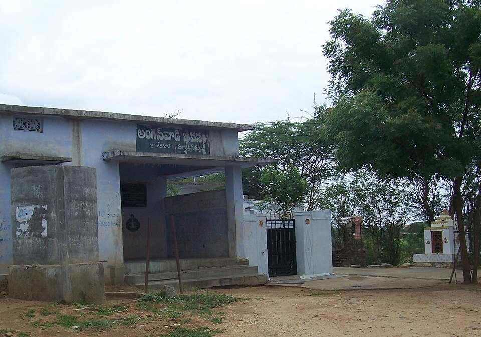 A single-storey pale blue village anganwadi building with a covered porch and a board in Telugu reading Anganwadi Bhavan above the entrance, on bare earth beside a neem tree.