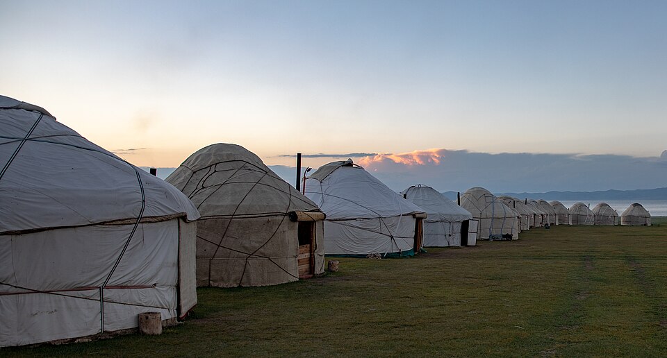 A row of white felt yurts with wooden door frames pitched on open green grassland at dusk, beside a lake with low hills on the far horizon
