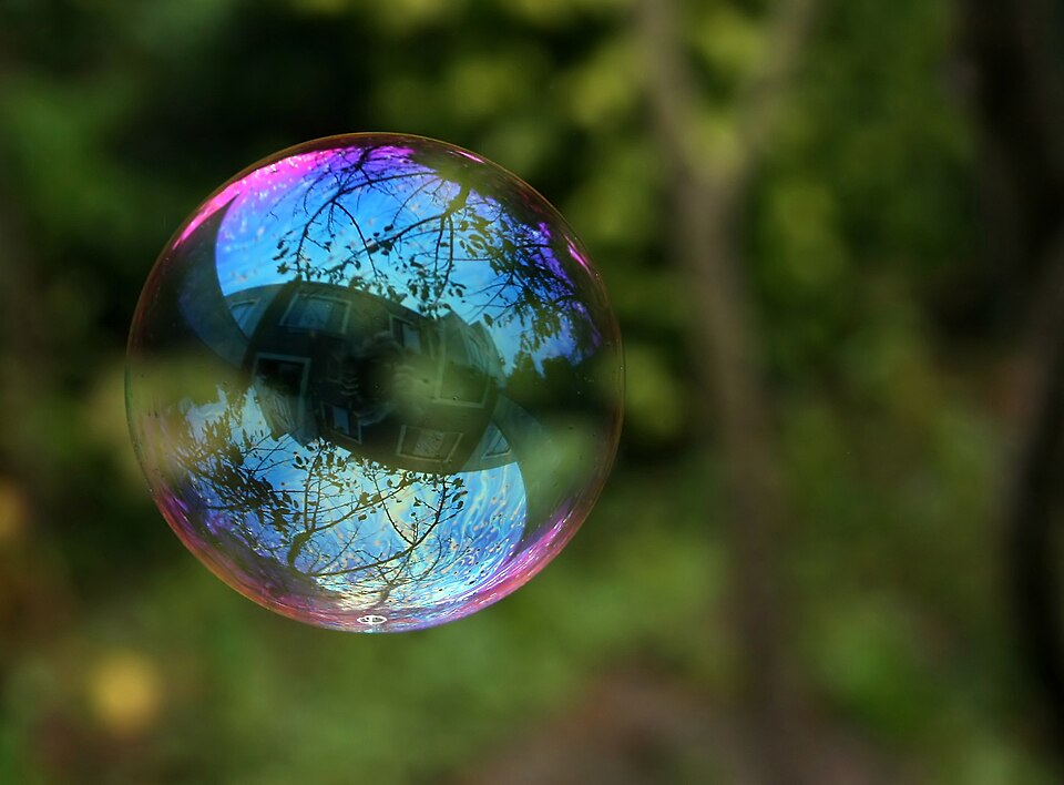 A single soap bubble floating in the air against blurred green foliage, its thin film shimmering with pink, blue and violet interference colours and mirroring trees and a house