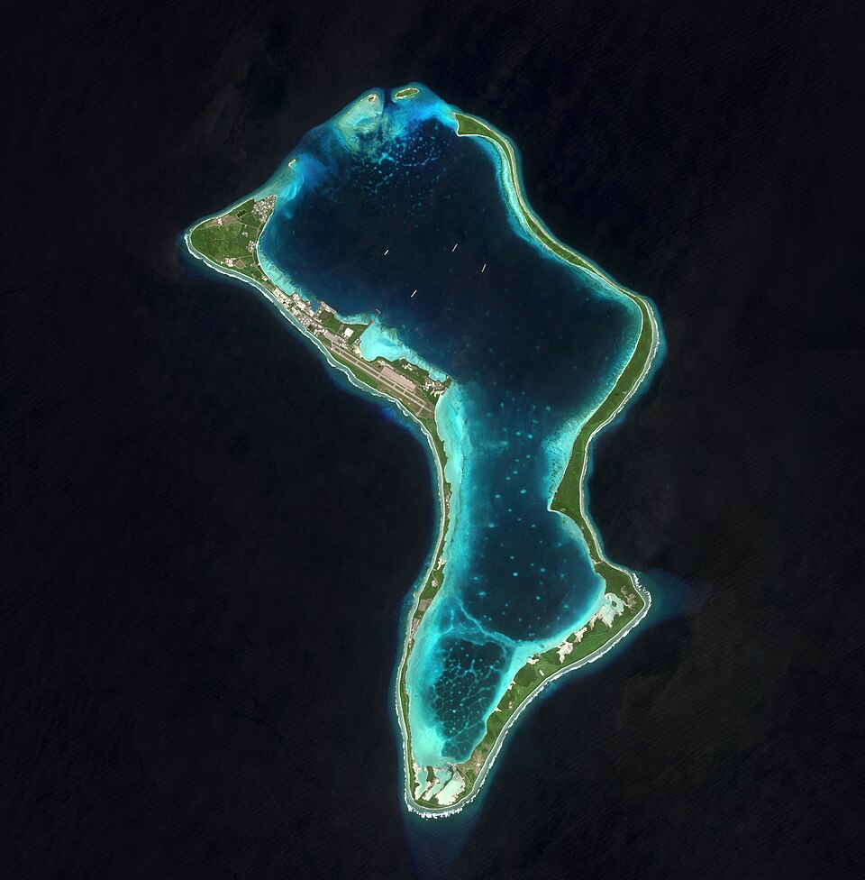 A satellite view of the V-shaped coral atoll of Diego Garcia — a narrow green rim of land enclosing a turquoise lagoon dotted with anchored ships, with an airstrip along its north-western arm and dark deep ocean all around