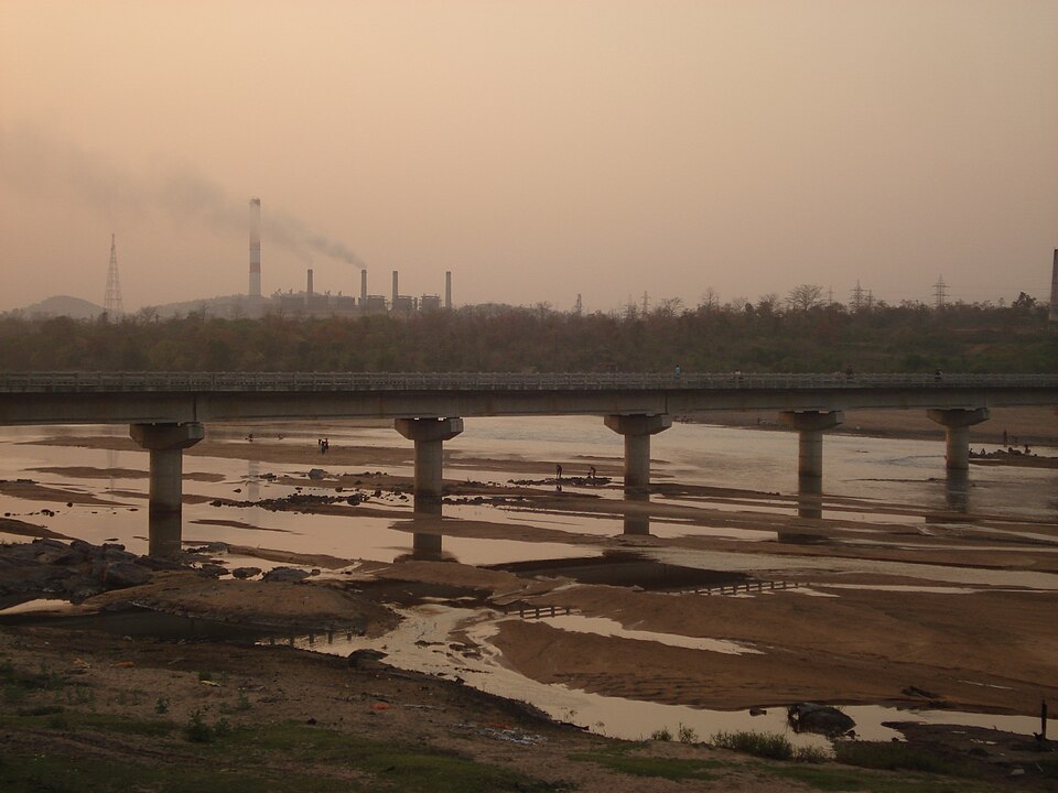 A broad sandy river bed of the Damodar with shallow braided channels, crossed by a concrete pillared road bridge, and the smoking chimneys of a coal-fired power station on the far bank in hazy evening light