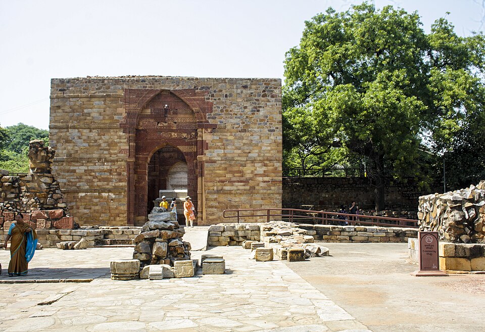 The square roofless stone chamber of Iltutmish tomb in the Qutb complex at Mehrauli, its doorway framed by a deeply carved red sandstone pointed arch, with loose carved blocks and a few visitors in the paved forecourt