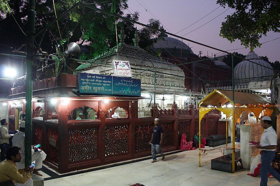 The red-painted, lattice-screened tomb pavilion of Amir Khusrau inside the Hazrat Nizamuddin dargah complex at dusk, its signboard naming him in Hindi and Urdu, with the domes of the dargah rising behind