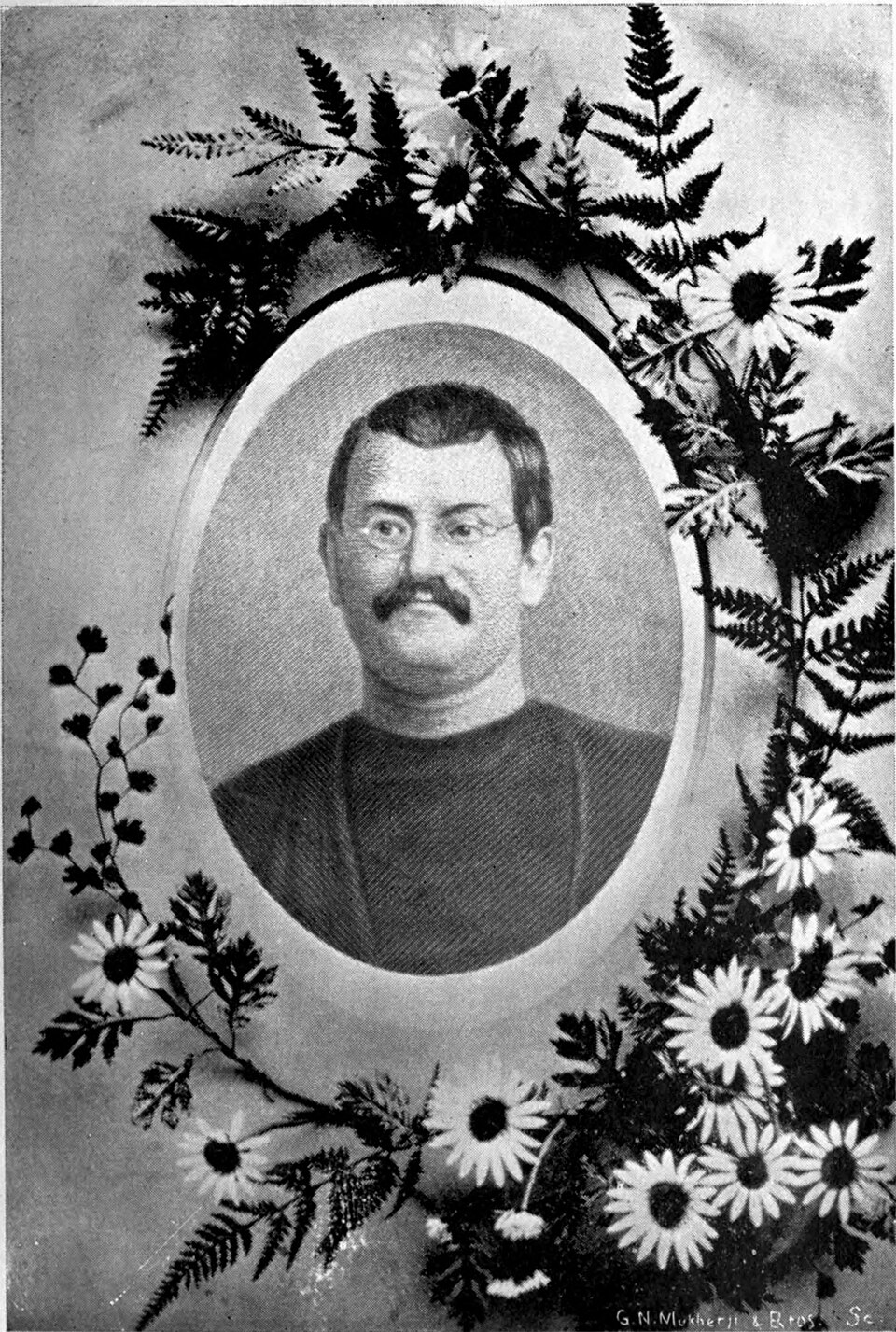 A nineteenth-century black-and-white oval portrait of Keshab Chandra Sen, a moustached man in round spectacles and a dark robe, set within a printed wreath of ferns and daisies