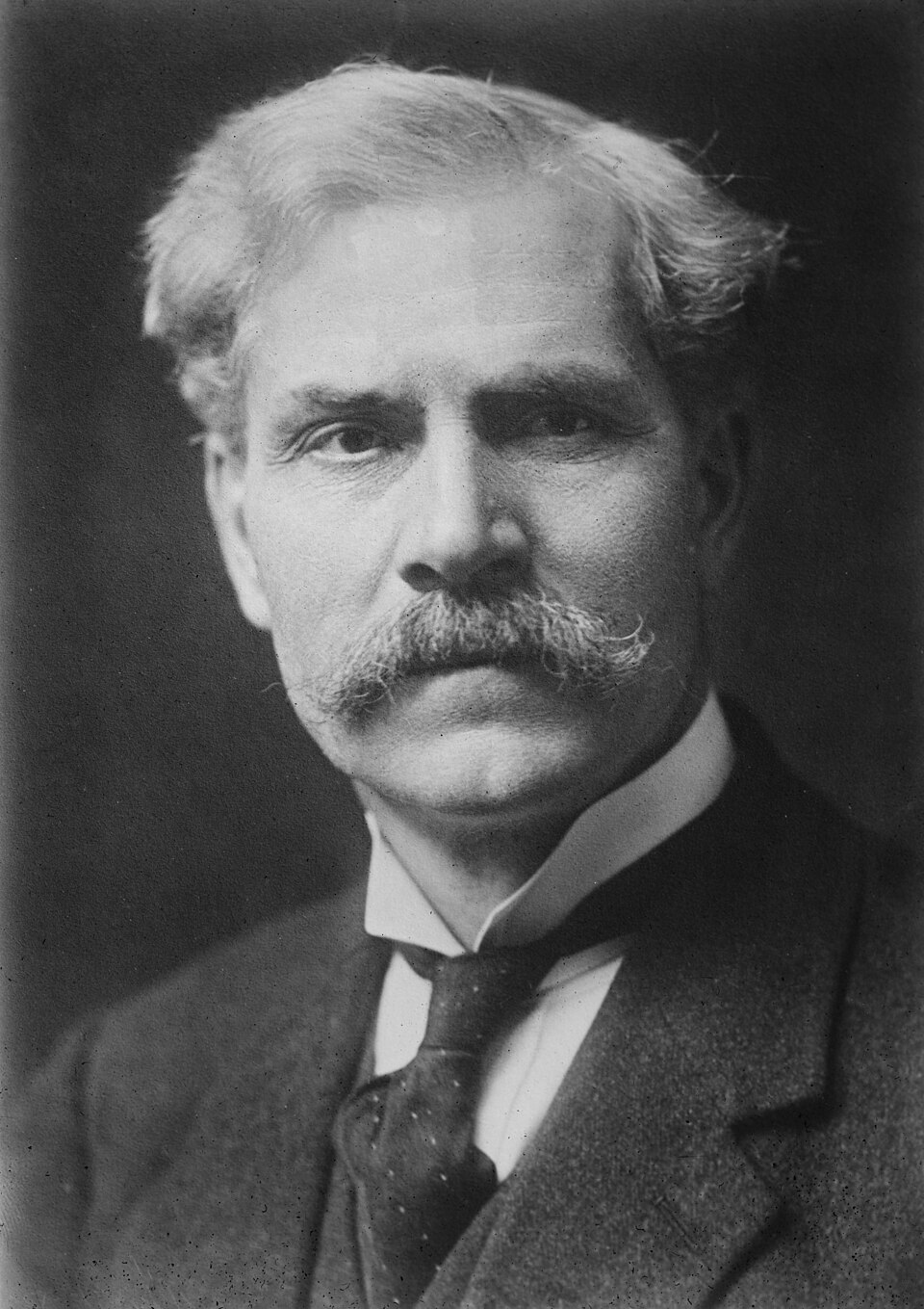 A black-and-white studio head-and-shoulders portrait of Ramsay MacDonald, an elderly man with swept-back white hair and a full white moustache, wearing a dark suit, waistcoat and tie