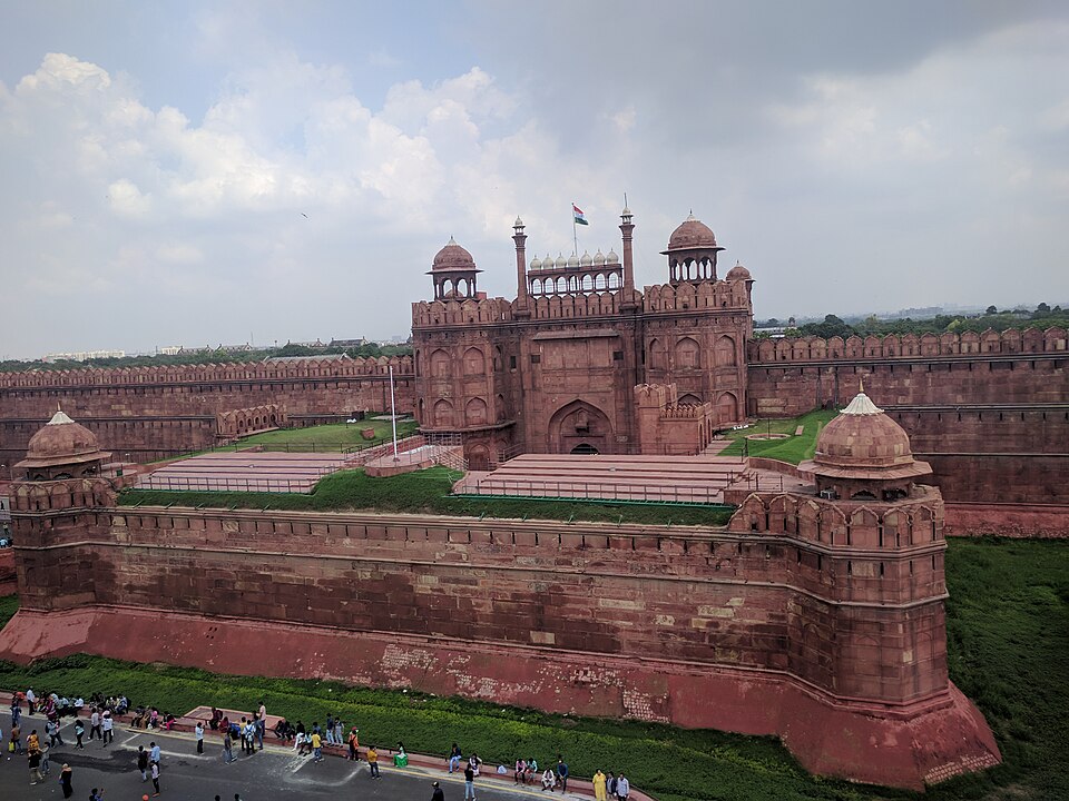 An elevated view of the Lahori Gate of the Red Fort in Delhi — red sandstone ramparts, domed pavilions and a barbican wall, with the Indian tricolour flying above the gate
