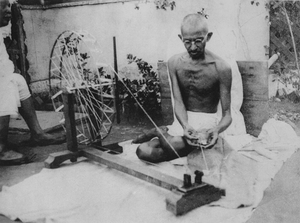 A historical black-and-white photograph of Mahatma Gandhi, bare-chested in a dhoti and round spectacles, sitting cross-legged on a white cloth and drawing thread from a large wooden charkha spinning wheel beside him